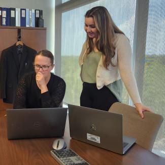 Two people in professional dress standing at a table looking a laptop.